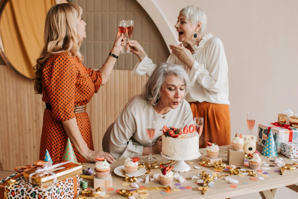 Three senior women celebrating a 60th birthday with cake and champagne indoors.